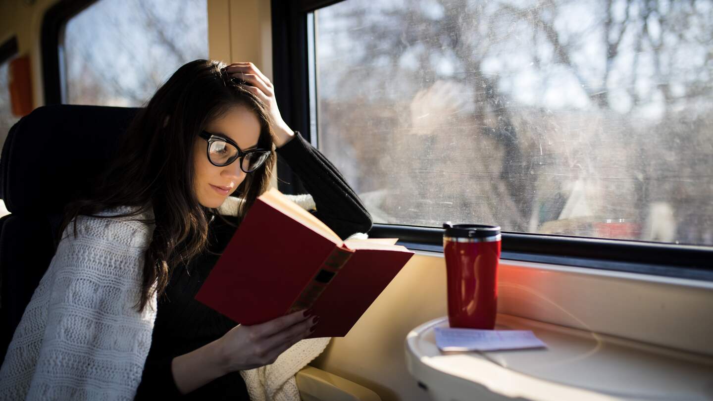 Entspannte Zugreise - Frau sitzt am Fensterplatz im Zug und genießt das Buch und den heißen Kaffee auf dem Tisch vor sich | © Gettyimages.com/DusanManic