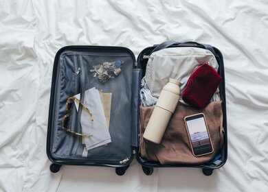 Ein geöffneter Koffer liegt auf dem Bett, gepackt für eine bevorstehende Reise. Darin befinden sich eine wiederverwendbare Wasserflasche, eine Kosmetiktasche, eine Sonnenbrille und Reisetickets. | © Gettyimages.com/FreshSplash
