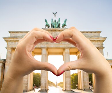 Hande mit rot lackierten Fingernägeln formen ein Herz vor dem Brandenburger Tor in Berlin  | © Gettyimages.com/Manuel-F-O