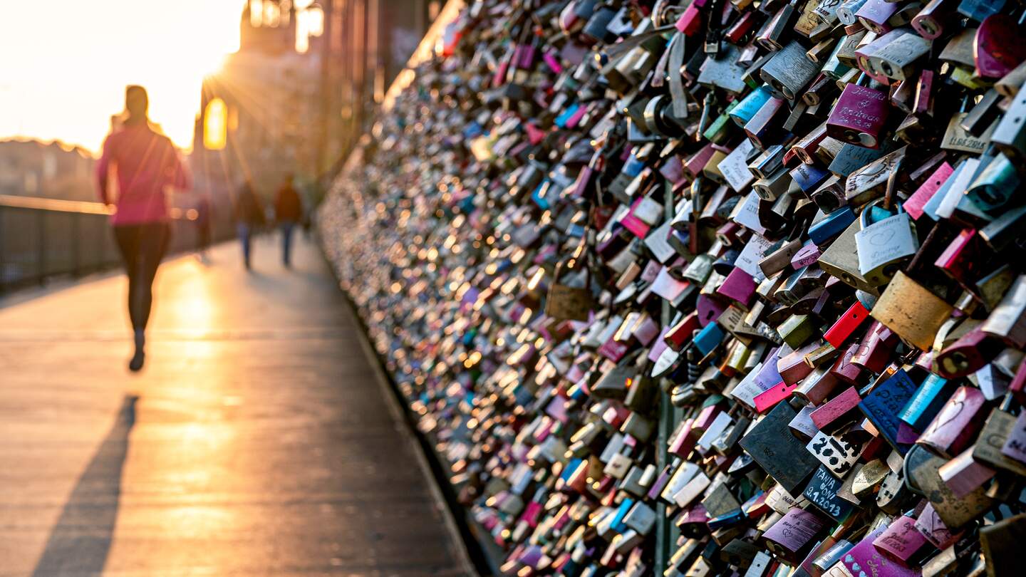 Liebesschlösser am Zaun der Hohenzollernbrücke in Köln im warmen Abendlicht. Menschen, die vorbeikommen. | © Gettyimages.com/Thomas Ramsauer