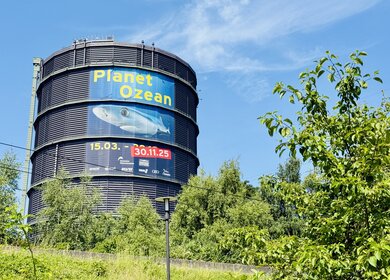 Großer, zylindrischer Gasometer mit Plakat zur Ausstellung ‚Planet Ozean‘ in gelber und weißer Schrift auf blauem Hintergrund. Das Plakat zeigt einen Fisch und informiert über die Laufzeit der Ausstellung vom 15. März bis zum 30. November 2025. Umgeben von Bäumen unter blauem Himmel. | © Michelle Khan