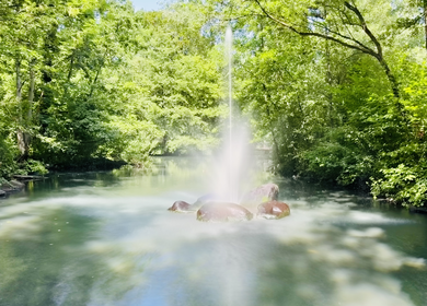 Ein Brunnen in Langzeitbelichtung in einem Park, umgeben von üppigen grünen Bäumen, der eine ruhige Atmosphäre im Freien schafft. | © Michelle Khan