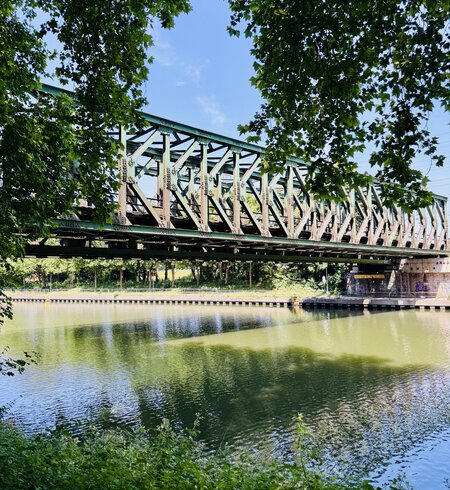Grüne Fachwerkbrücke aus Metall überquert einen Kanal, umgeben von Bäumen. Das Wasser spiegelt die Brücke und das umliegende Grün. Die Brücke ruht auf steinernen Widerlagern, der Himmel ist blau mit etwas Licht. | © Michelle Khan