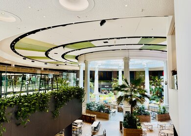 Innenaufnahme des Bereichs Coca-Cola Oasis im Westfield Centro in Oberhausen. Zu sehen ist ein modern gestalteter Gastronomiebereich mit runden Deckenelementen, weißen Säulen und tropischen Pflanzen wie Palmen. Sitzbereiche auf zwei Etagen, durchzogen von viel Grün und natürlichem Licht. | © Michelle Khan
