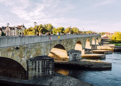 Blick auf die historische Steinbruecke in Regensburg, Bayern, Deutschland. Touristen laufen unter strahlend blauem Himmel ueber die Bruecke. | © Gettyimages.com/Nikada