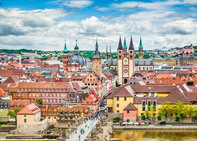  Panoramaansicht der Stadt Wuerzburg in Deutschland, mit einer Bruecke ueber einen Fluss, die zu einem Bereich mit verschiedenen historischen Gebaeuden fuehrt, darunter markante Kirchtuerme und Tuerme. | © Gettyimages.com/JR Photography