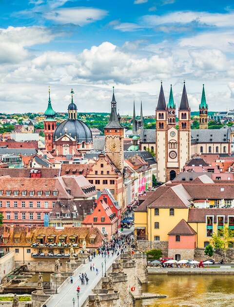  Panoramaansicht der Stadt Wuerzburg in Deutschland, mit einer Bruecke ueber einen Fluss, die zu einem Bereich mit verschiedenen historischen Gebaeuden fuehrt, darunter markante Kirchtuerme und Tuerme. | © Gettyimages.com/JR Photography