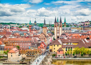  Panoramaansicht der Stadt Wuerzburg in Deutschland, mit einer Bruecke ueber einen Fluss, die zu einem Bereich mit verschiedenen historischen Gebaeuden fuehrt, darunter markante Kirchtuerme und Tuerme. | © Gettyimages.com/JR Photography