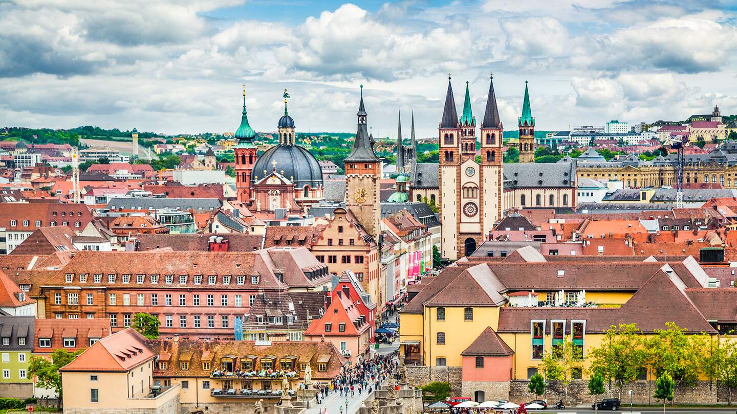  Panoramaansicht der Stadt Wuerzburg in Deutschland, mit einer Bruecke ueber einen Fluss, die zu einem Bereich mit verschiedenen historischen Gebaeuden fuehrt, darunter markante Kirchtuerme und Tuerme. | © Gettyimages.com/JR Photography