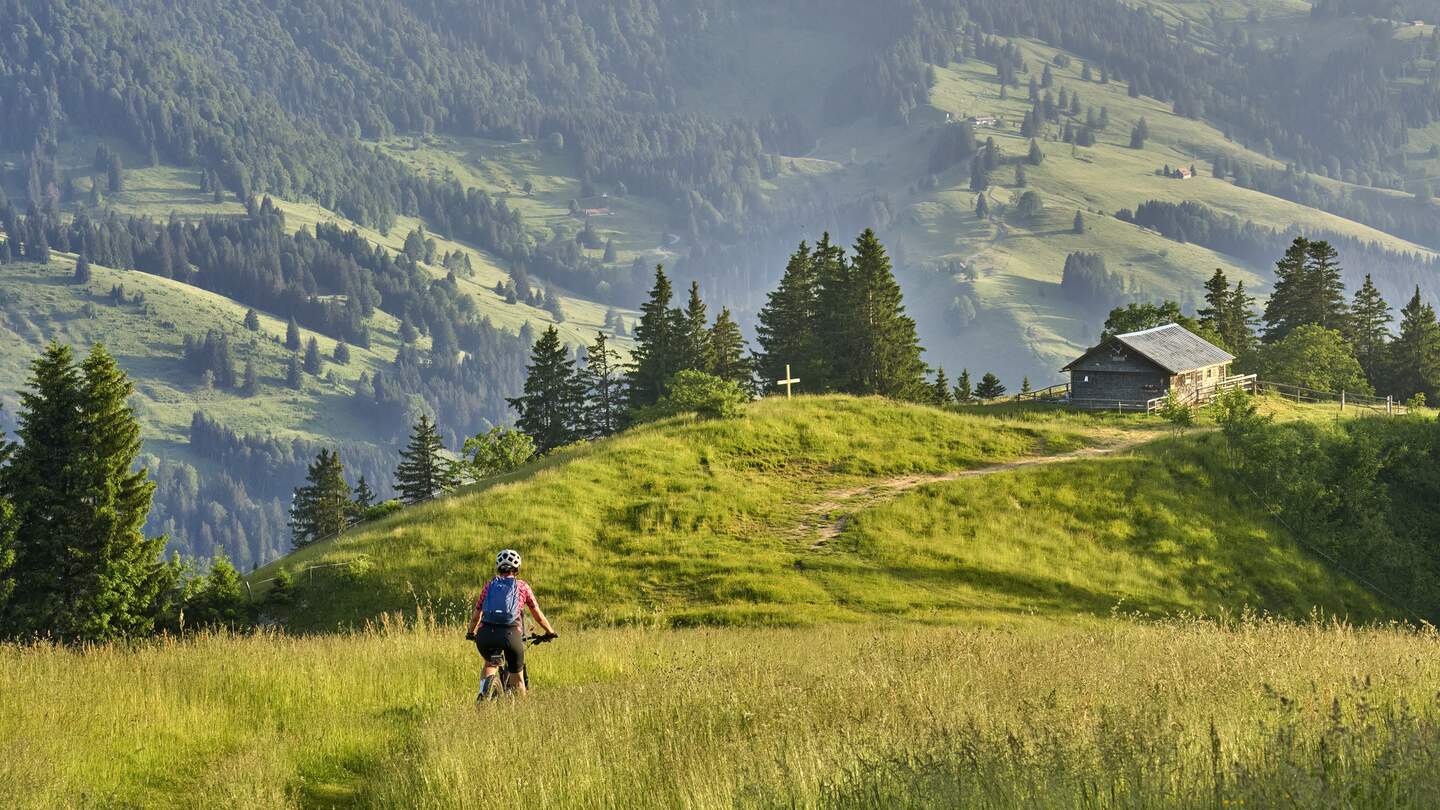 Frau faehrt ihr Elektro-Mountainbike in den Bergen oberhalb von Oberstaufen mit Nagelfluh-Bergkette im Hintergrund, Allgaeuer Alpen | © Gettyimages.com/Uwe Moser