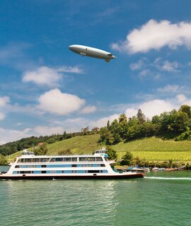 Passagierfähre von Meersburg auf ihrer Route über den Bodensee an einem Sommertsg, mit Weinbergen und einem Zeppelin im Hintergrund | ©  Gettyimages.com/olgysha2008