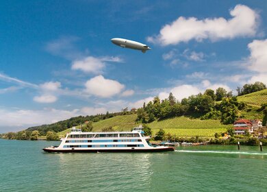 Passagierfähre von Meersburg auf ihrer Route über den Bodensee an einem Sommertsg, mit Weinbergen und einem Zeppelin im Hintergrund | ©  Gettyimages.com/olgysha2008