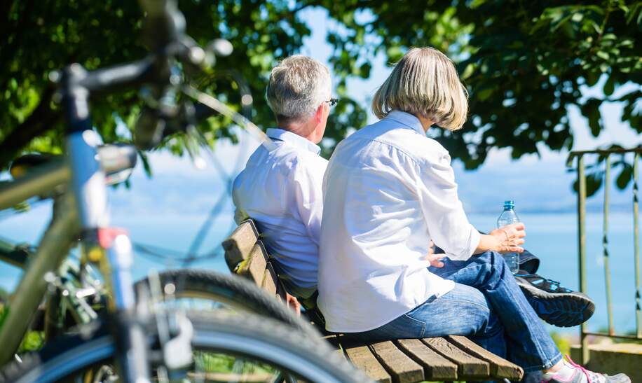Bestager-Paar genießt bei einer Fahrradtour-Auszeit auf einer Bank die Aussicht auf den Bodensee | © Gettyimages.com/kzenon