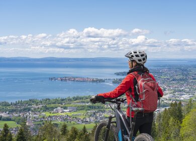 Frau mit Elektro-Mountainbike steht auf einem Berg und schaut hinunter auf das Panorama des Bodensees | © GettyImages.com/Uwe Moser