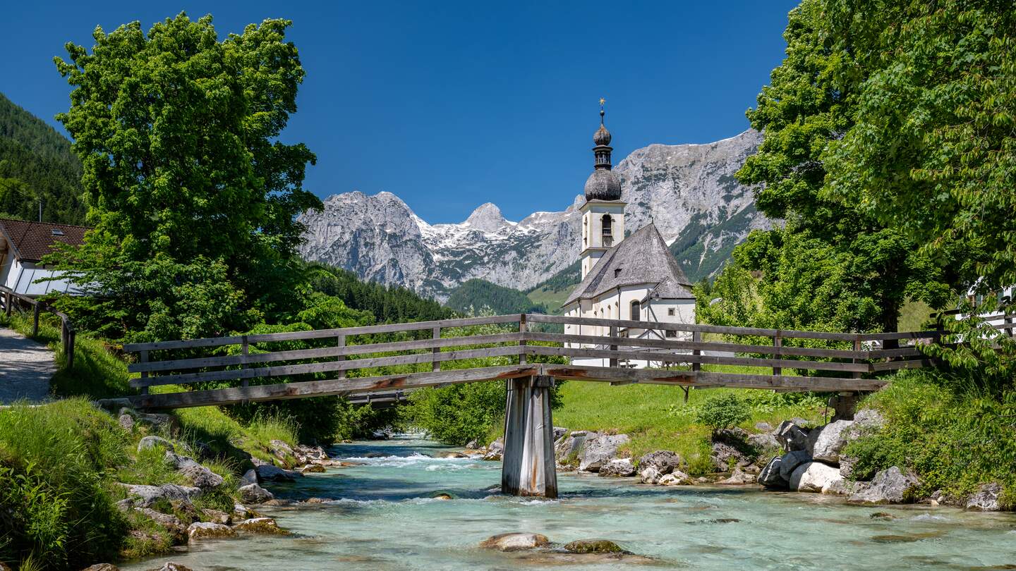 Blick auf die idyllische Kirche St. Sebastian in Ramsau im Sommer, Holzbruecke fuehrt ueber einen Bergbach, Bayern, Deutschland | © Gettyimages.com/auerimages