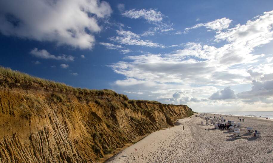Links sieht man eine steile Klippe. Rechts Strandkoerbe und Menschen am Strand | © Gettyimages.com/viktorkunz