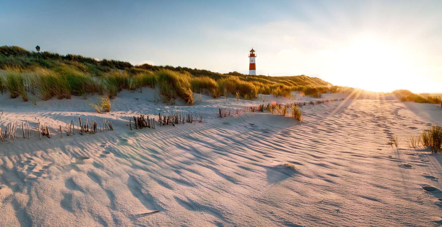 Ein rot weisser Leuchtturm in den Duenen, die Sonne geht unter und taucht die Grashalme auf den Duenen in goldfarbenes Licht | © Gettyimages.com/mthaler