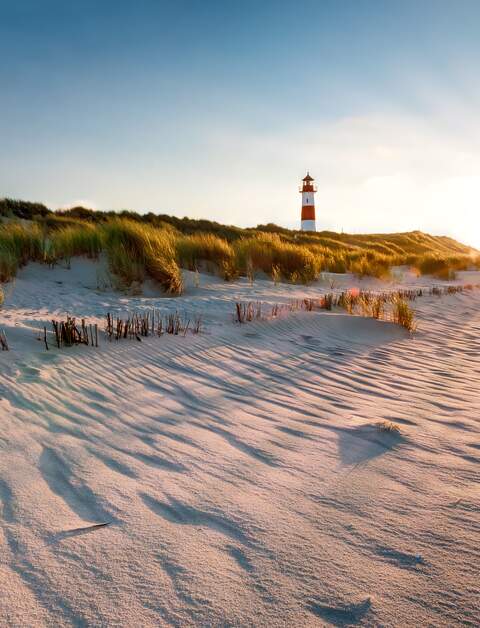 Ein rot weisser Leuchtturm in den Duenen, die Sonne geht unter und taucht die Grashalme auf den Duenen in goldfarbenes Licht | © Gettyimages.com/mthaler