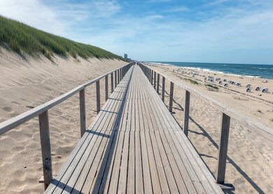 Die bekannte Strandpromenade mit dem Holzsteg in Richtung der Stadt Westerland, Hauptstadt der deutschen Insel Sylt in der Nordsee. Rechts der Strand voll mit Sttr4andkörben und Menschen, links die Düne | © Gettyimages.com/karlhendriktittel