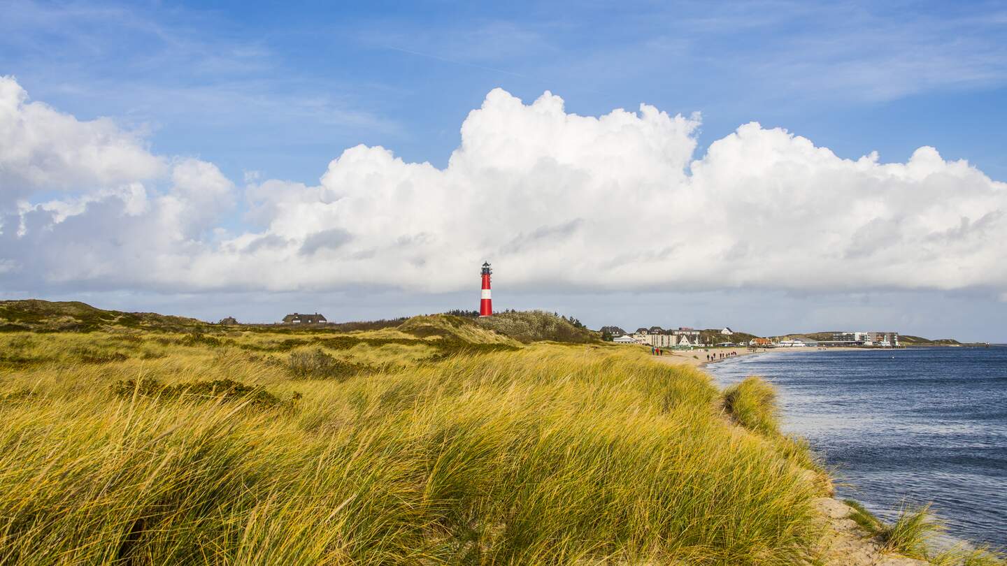 Der rot, weisse Leuchtturm ragt in den Duenen empor. Im Hintergrund sieht man den Strand mit Menschen und ein paar Haeuser: Rechts ist das Meer. | © Gettyimages/marcstephan