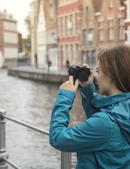 Weibliche Touristin fotografiert in Bruegge an einem Kanal, in dem ein Boot faehrt. | © Gettyimages.com/GoodLifeStudio