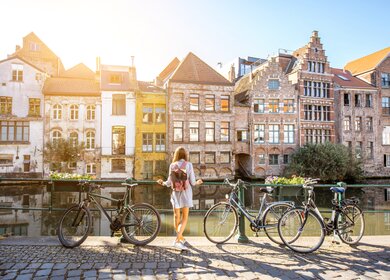 Eine Frau steht in Gent, Belgien an einem Fluss und blickt auf die Altstadt. Neben ihr stehen Fahrräder | © Gettyimages.com/RossHelen