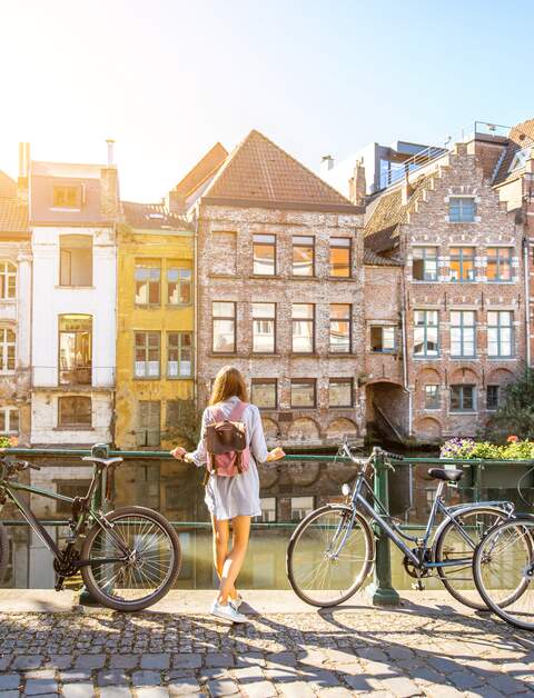 Eine Frau steht in Gent, Belgien an einem Fluss und blickt auf die Altstadt. Neben ihr stehen Fahrräder | © Gettyimages.com/RossHelen