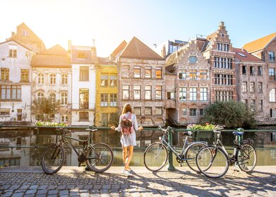 Eine Frau steht in Gent, Belgien an einem Fluss und blickt auf die Altstadt. Neben ihr stehen Fahrräder | © Gettyimages.com/RossHelen