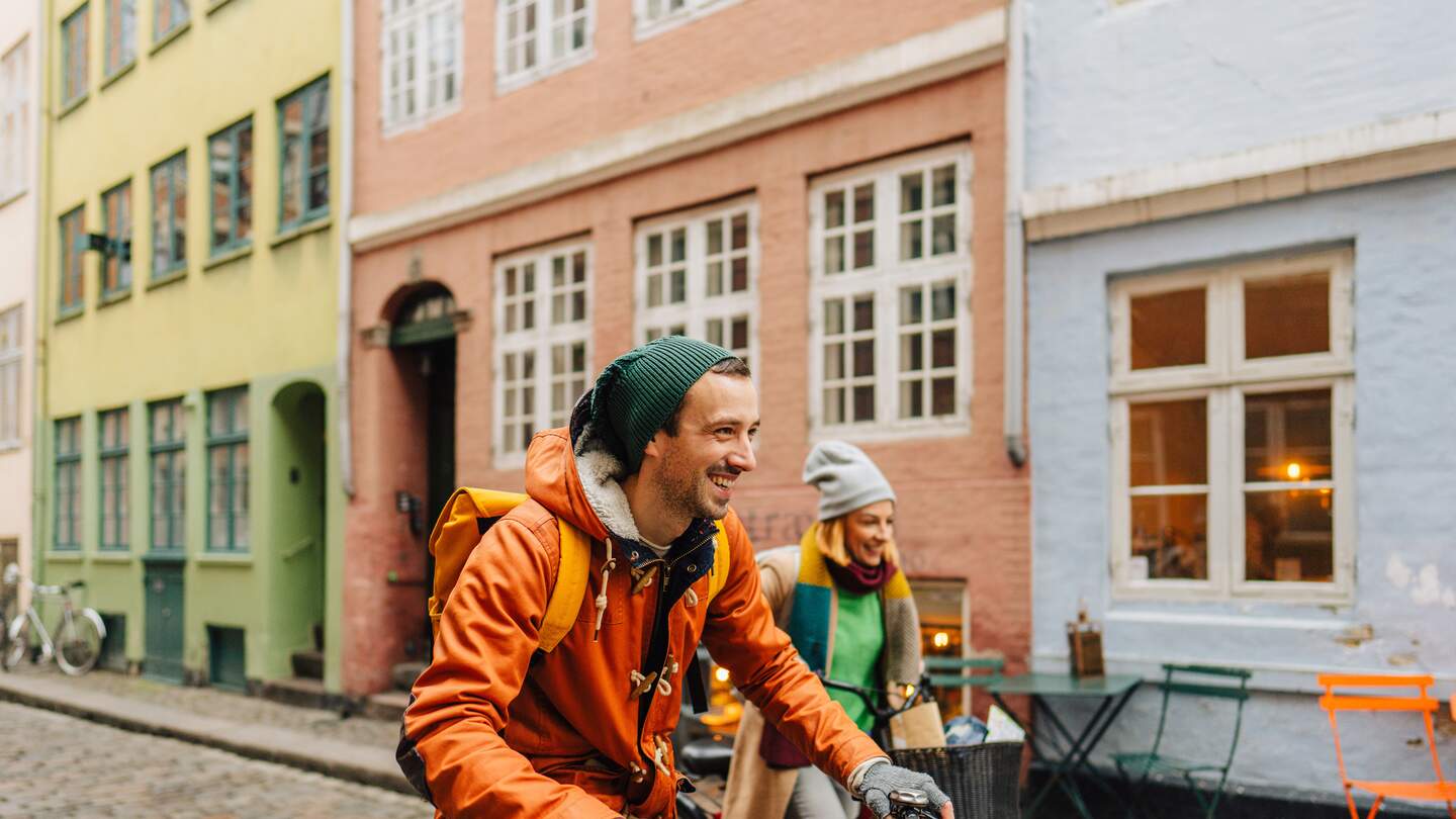 Paar faehrt mit dicken Jacken und Muetzen auf Fahrraedern durch die Innenstadt in Kopenhagen, Daenemark | © GettyImages.com/AleksandarNakic
