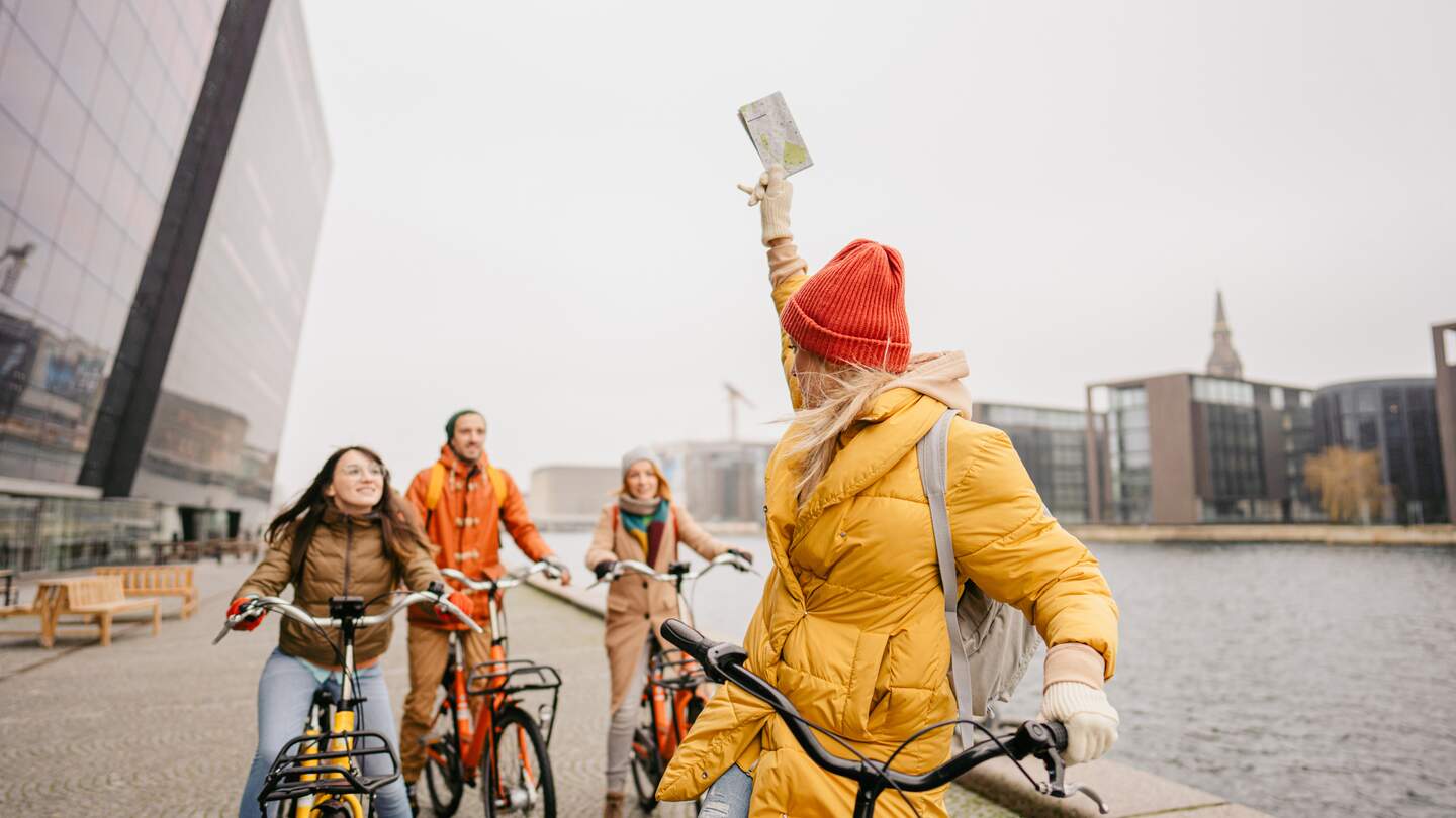 Gruppe Touristen mit dicken Jacken und Muetzen auf Fahrrädern am Hafen in Kopenhagen, Dänemark, vor der Det Kongelige Bibliothek | © GettyImages.com/AleksandarNakic