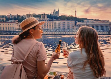 Zwei glueckliche Maedchen trinken Wein aus Flaschen und beobachten eine Sonne geht ueber Saone Fluss in Lyon  | © Gettyimages.com/frantic00