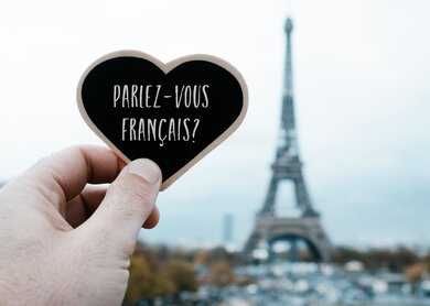 Nahaufnahme einer Hand, die ein herzfoermiges Schild mit der Frage haelt, ob Sie Franzoesisch sprechen, das darin auf Franzoesisch geschrieben ist, vor dem Eiffelturm in Paris, Frankreich | © Gettyimages.com/nito100