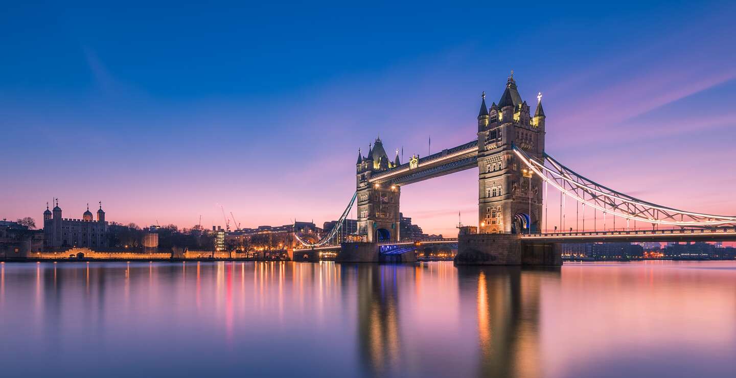 Tower Bridge in London bei blauem und rosa Abendhimmel, mit beleuchteten Türmen und ruhigem Wasser, das die Brücke und Lichter reflektiert. | © Gettyimages.com/	Trung Pham