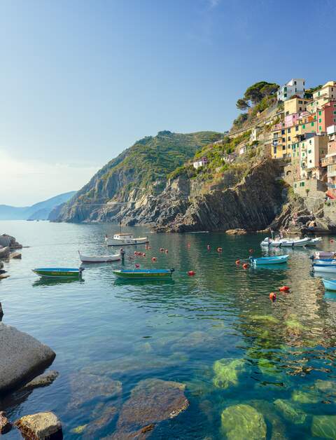 Idyllischer Blick vom smaragdgruenen Mittelmeer auf die Stadt Riomaggiore im Cinque Terre Nationalpark.  Die bunten Häuser des Dorfes und der Hafen strahlen im Sonnenlicht, während kleine Fischerboote auf dem Wasser vor der Küste treiben. | © Gettyimages.com/spooh