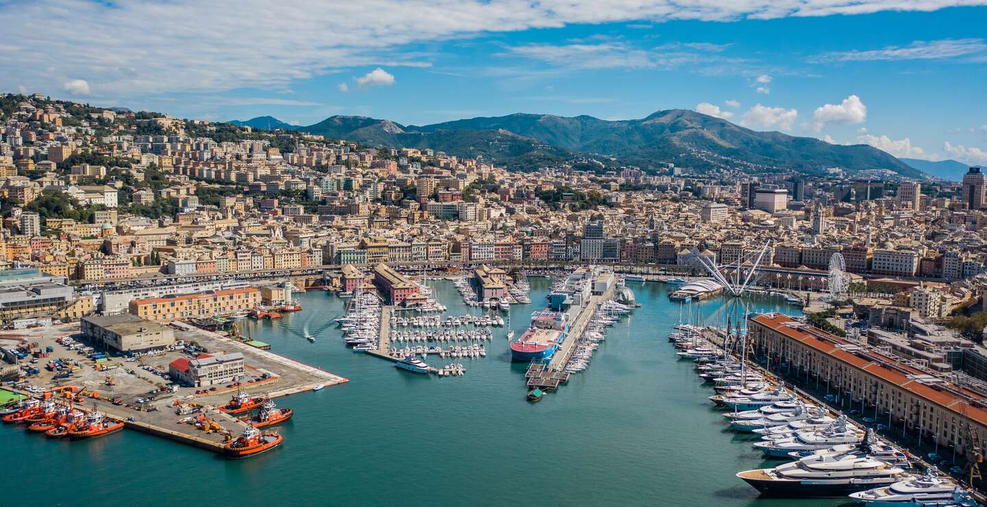 Luftaufnahme von Genua vom Meer aus, mit dem Hafen für Boote und Yachten, dem Riesenrad und der Stadt im Vordergrund, umgeben von grünen Hügeln im Hintergrund. | © Gettyimages.com/Medvedkov