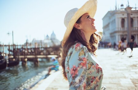 Glueckliche Touristin im Blumenkleid mit Hut, die an einem sonnigen Tag in Venedig am Ufer flaniert.  | © Gettyimages.com/CentralITAlliance