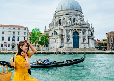 Frau im gelben Sonnenkleid sitzt am Ufer mit Blick auf den grossen Kanal in Venedig. | © Gettyimages.com/Vera_Petrunina