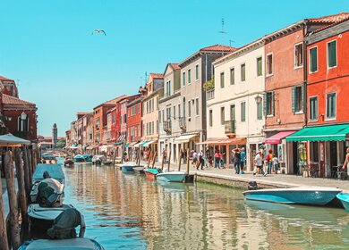Bunte Haeuser auf der Insel Murano, die an einem kleinen Kanal stehen. Touristen flanieren entlang des Kanals an einem sonnigen Tag. | © Gettyimages.com/Gavin Guan