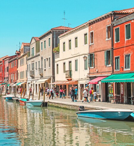 Bunte Haeuser auf der Insel Murano, die an einem kleinen Kanal stehen. Touristen flanieren entlang des Kanals an einem sonnigen Tag. | © Gettyimages.com/Gavin Guan