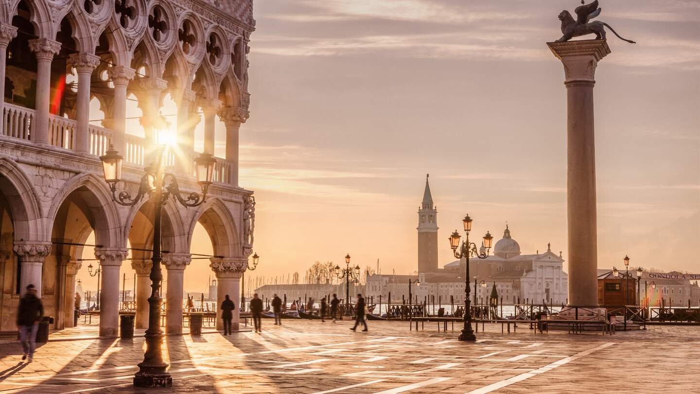 Auf dem Markusplatz in Venedig laufen Menschen. Die Sonne scheint durch den Dogenpalast. | © Gettyimages.com/tunart