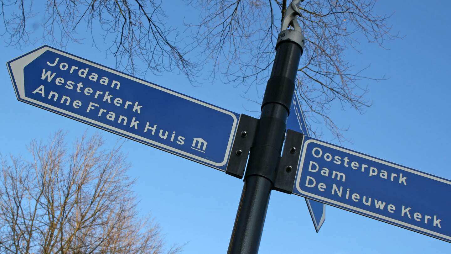 einen Wegweiser in Amsterdam mit blauen Schildern, die zu verschiedenen Sehenswuerdigkeiten wie dem Anne Frank Haus und dem Dam fuehren. | ©  Gettyimages.com/Lya_Cattel