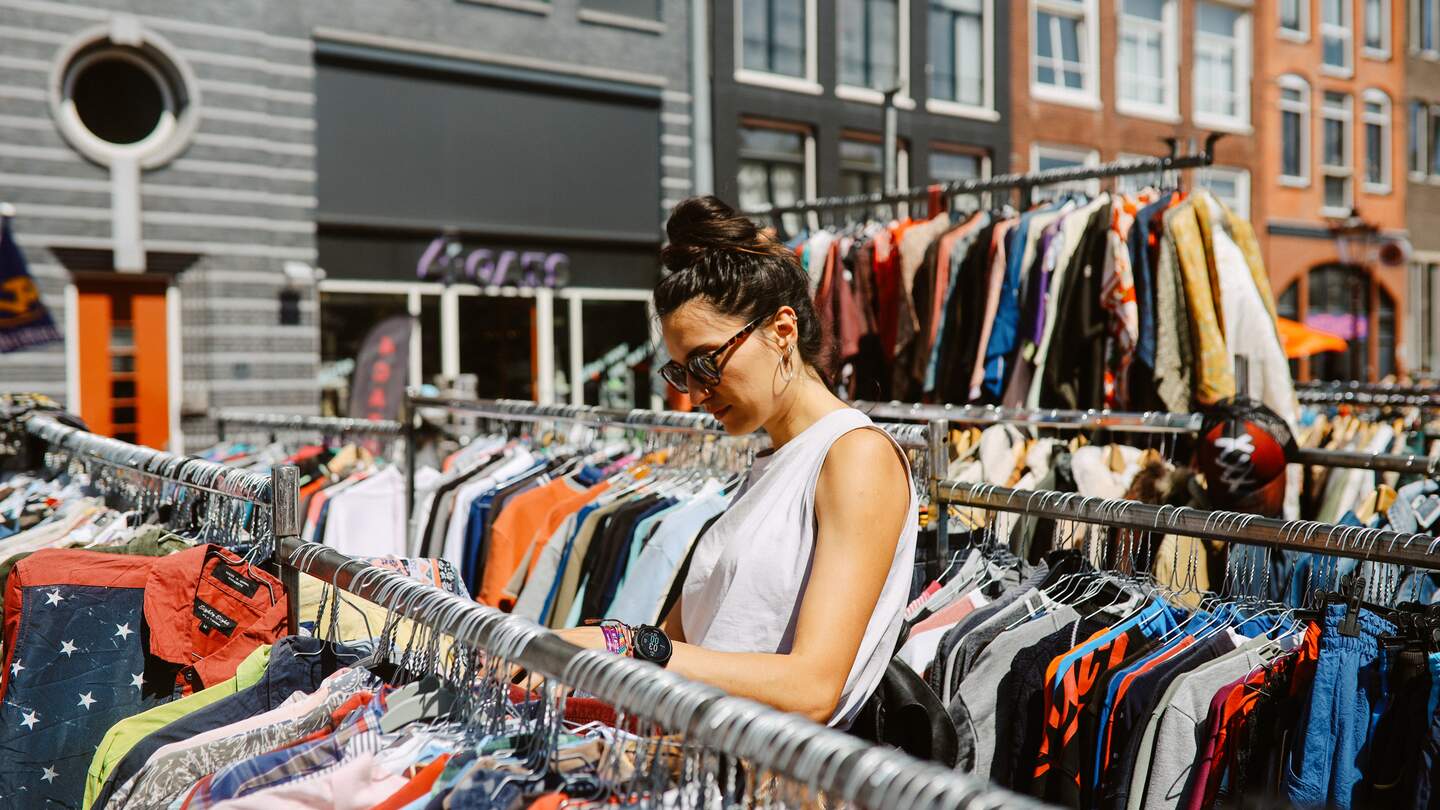 Frau mit Sonnenbrille und ärmelloser Bluse schaut durch die Kleiderstaender eines Flohmarktes in Amsterdam | © Gettyimages.com/lechatnoir
