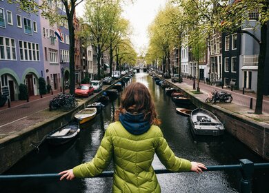 Junge Frau in hellgruener, taillerter Steppjacke steht auf einer Grachtenbrücke in Amsterdam und blickt auf den Kanal | © Gettyimages.com/bortnikau