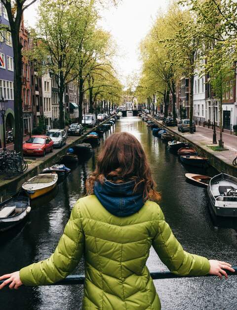 Junge Frau in hellgruener, taillerter Steppjacke steht auf einer Grachtenbrücke in Amsterdam und blickt auf den Kanal | © Gettyimages.com/bortnikau