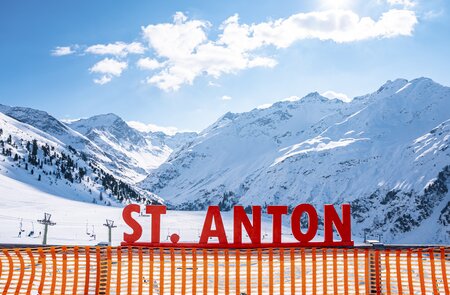 Großer Schriftzug „St. Anton“ auf einem Geländer im Skigebiet, an einem sonnigen Tag mit verschneiten Bergen im Hintergrund. | © Gettyimages.com/AlexKane