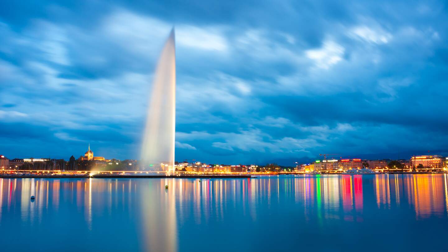 Genfer Fontaene im Genfersee am Abend mit beleuchteten Haeusern | © Gettyimages.com/xenotar