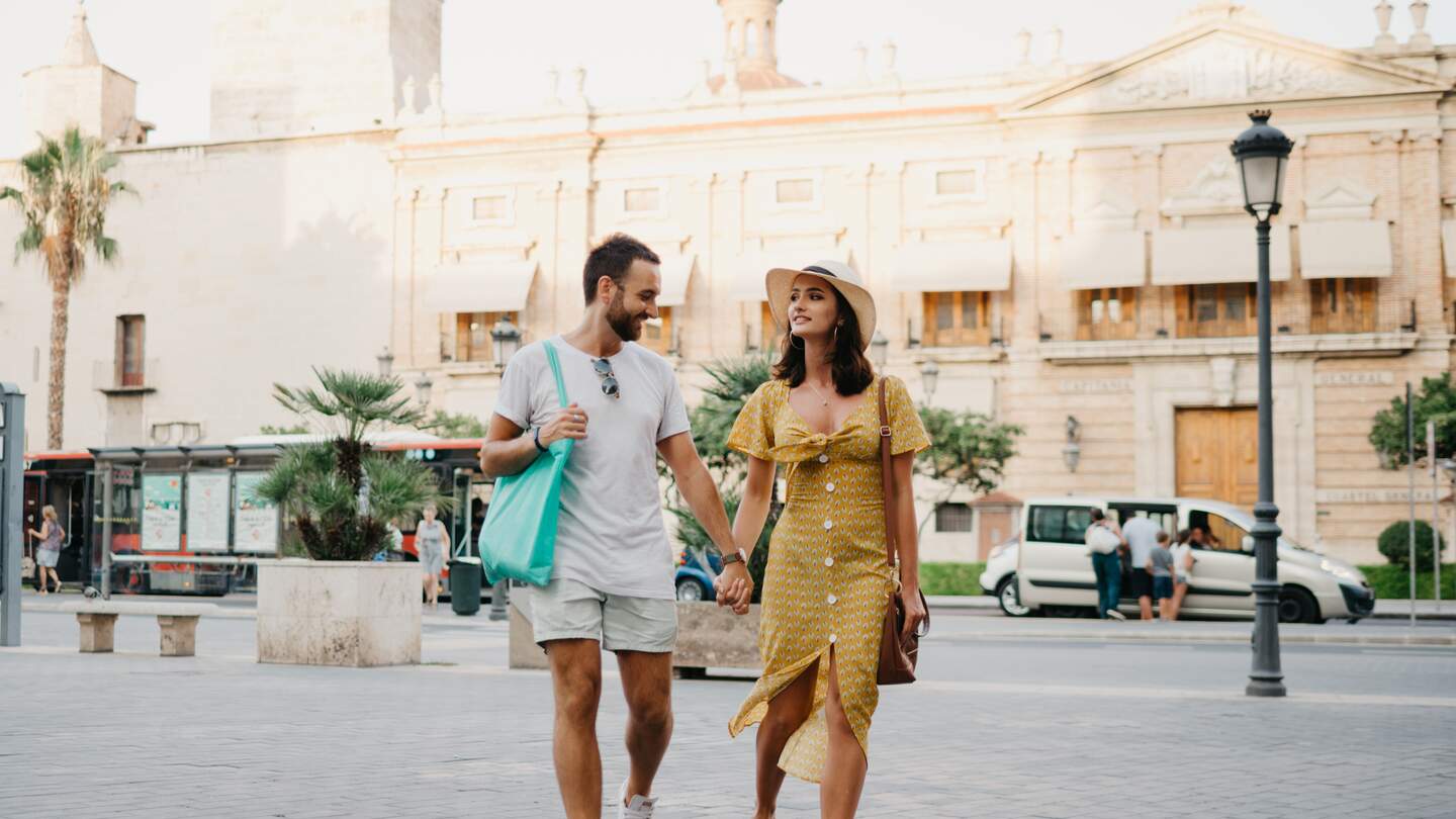 Mädchen mit Hut und Kleid und ihr Freund mit Bart und Sonnenbrille entspannen in der Altstadt und halten sich an der Hand. Ein paar Touristen beim Sonnenuntergang in Valencia. | © Gettyimages.com/Tyukin