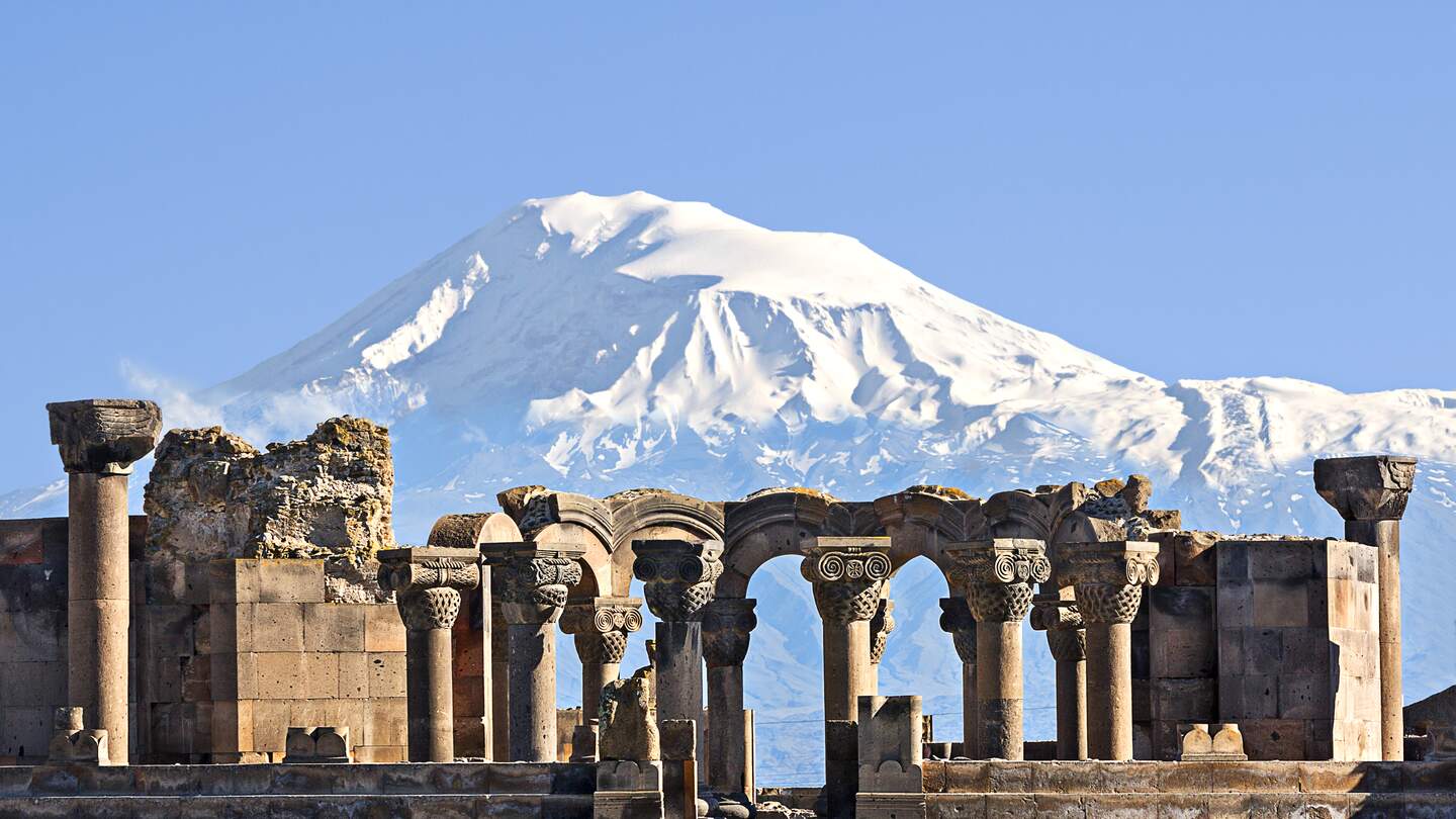 Antike Steinruinen vor einem schneebedeckten Bergmassiv unter klarem Himmel; beeindruckende Landschaftskulisse mit historischer Architektur und Natur. | © Gettyimages.com/Ozbalci