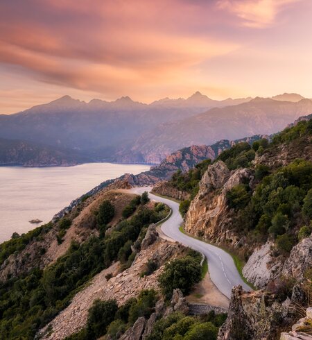 Die Strasse D824 schlaengelt sich von Capu Rossu entlang der Kueste in Richtung Piana an der Westkueste Korsikas, waehrend die fruehe Morgensonne die fernen Berge beleuchtet | © Gettyimages.com/77joningall