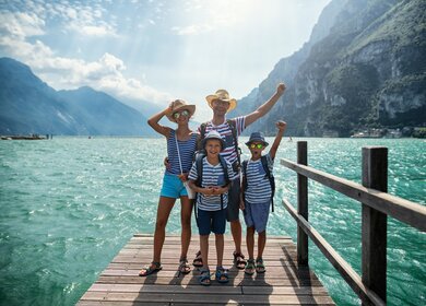 Familie geniesst Urlaub in Italien. Vater und Kinder stehen auf einem Pier in Riva del Garda und jubeln in die Kamera. Dahinter bietet sich ein herrlicher Blick auf den Gardasee, umgeben von den Alpen | © Gettyimages.com/Imgorthand